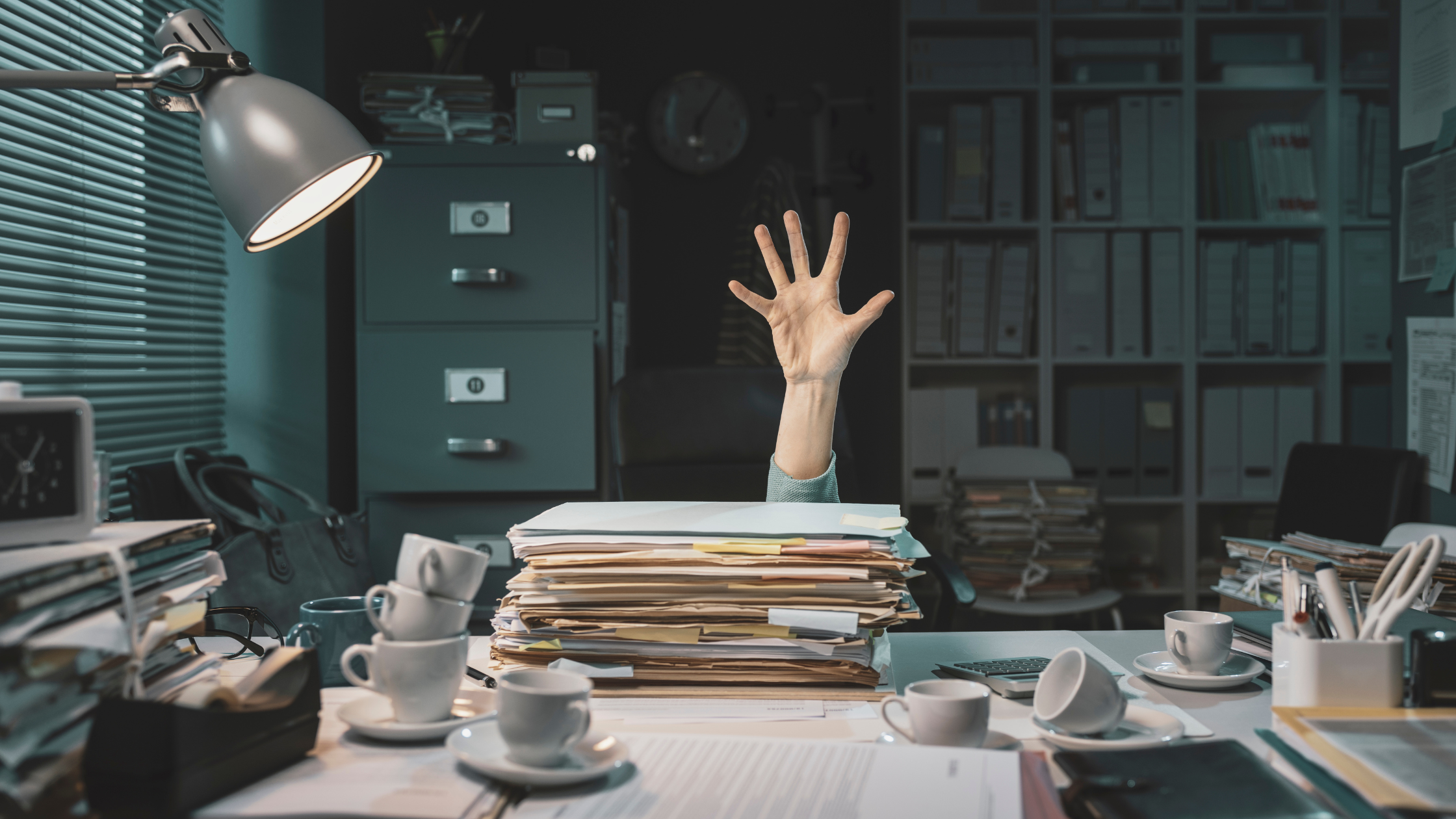 A hand reaching out from a large stack of paperwork on a cluttered office desk.